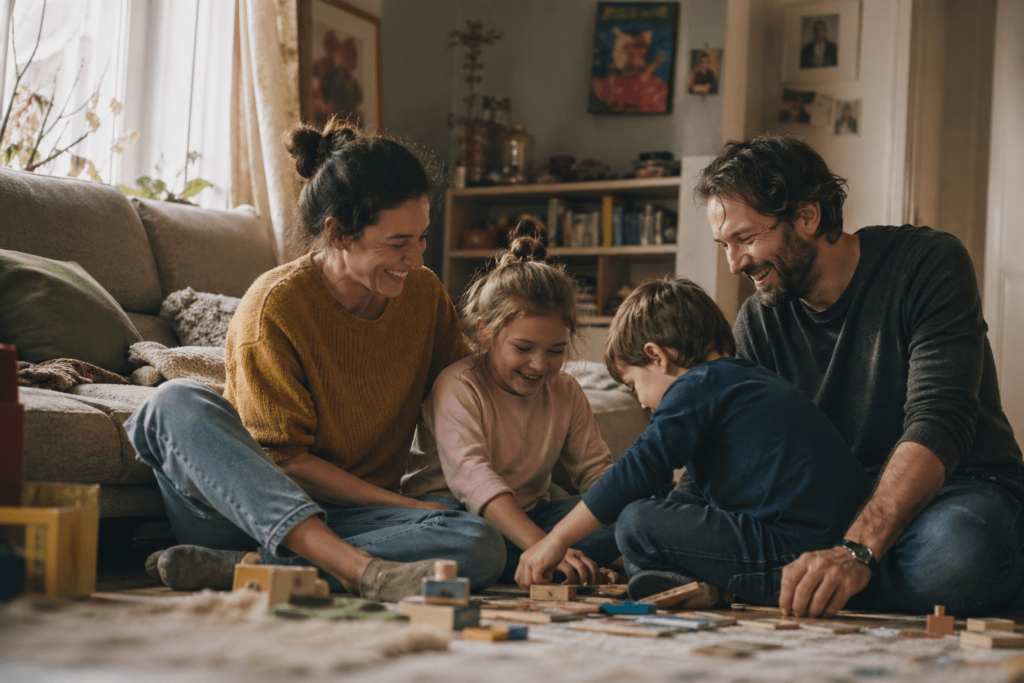A warm, candid photograph of a family of four sitting on a living room floor. A mother, father, and two young children are laughing and playing with wooden blocks. The lighting is soft and natural, creating a cozy, non-commercial atmosphere.