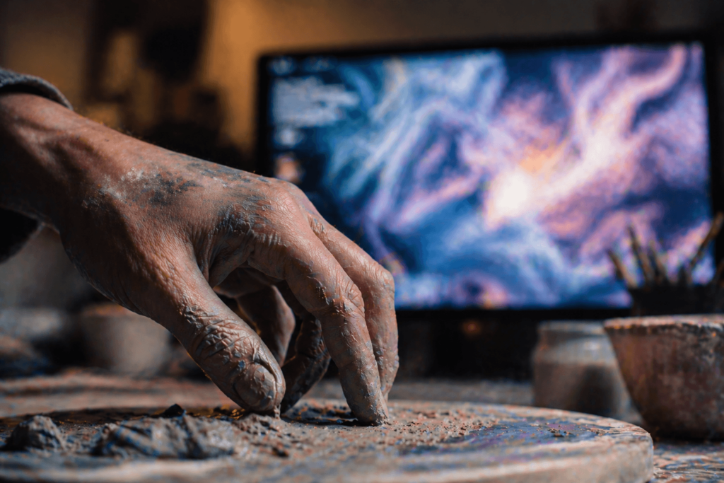 A close-up of a dirty, weathered human hand working with physical clay or paint in a workshop. In the blurred background, a computer monitor glows with complex AI-generated digital art, symbolizing the irreplaceable value of physical human touch in a digital age.