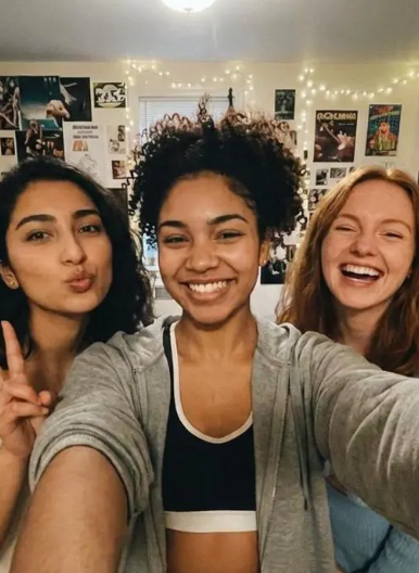 A close-up selfie of three happy young diverse women posing together indoors in a room decorated with posters and string lights.