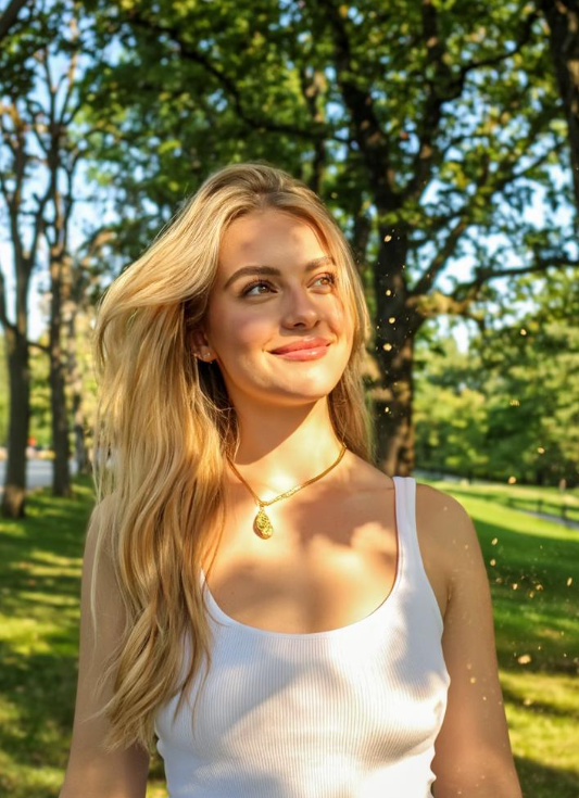 A natural photo of a smiling blonde woman in a park with dapple light, wearing a simple white ribbed tank top.