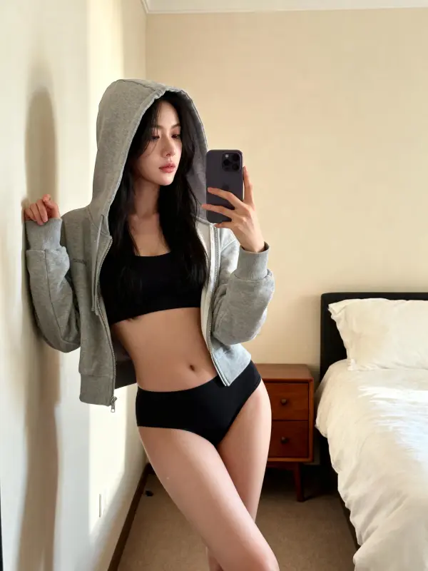 Realistic AI-generated woman in a grey zip-up hoodie and black sports bra taking a mirror selfie in a bedroom.