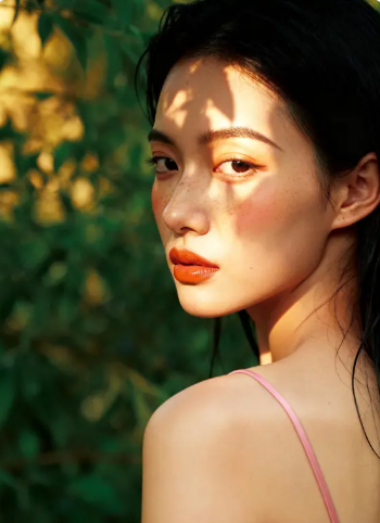 High-detail close-up of a woman with freckles and orange lipstick under dappled tree shadows.