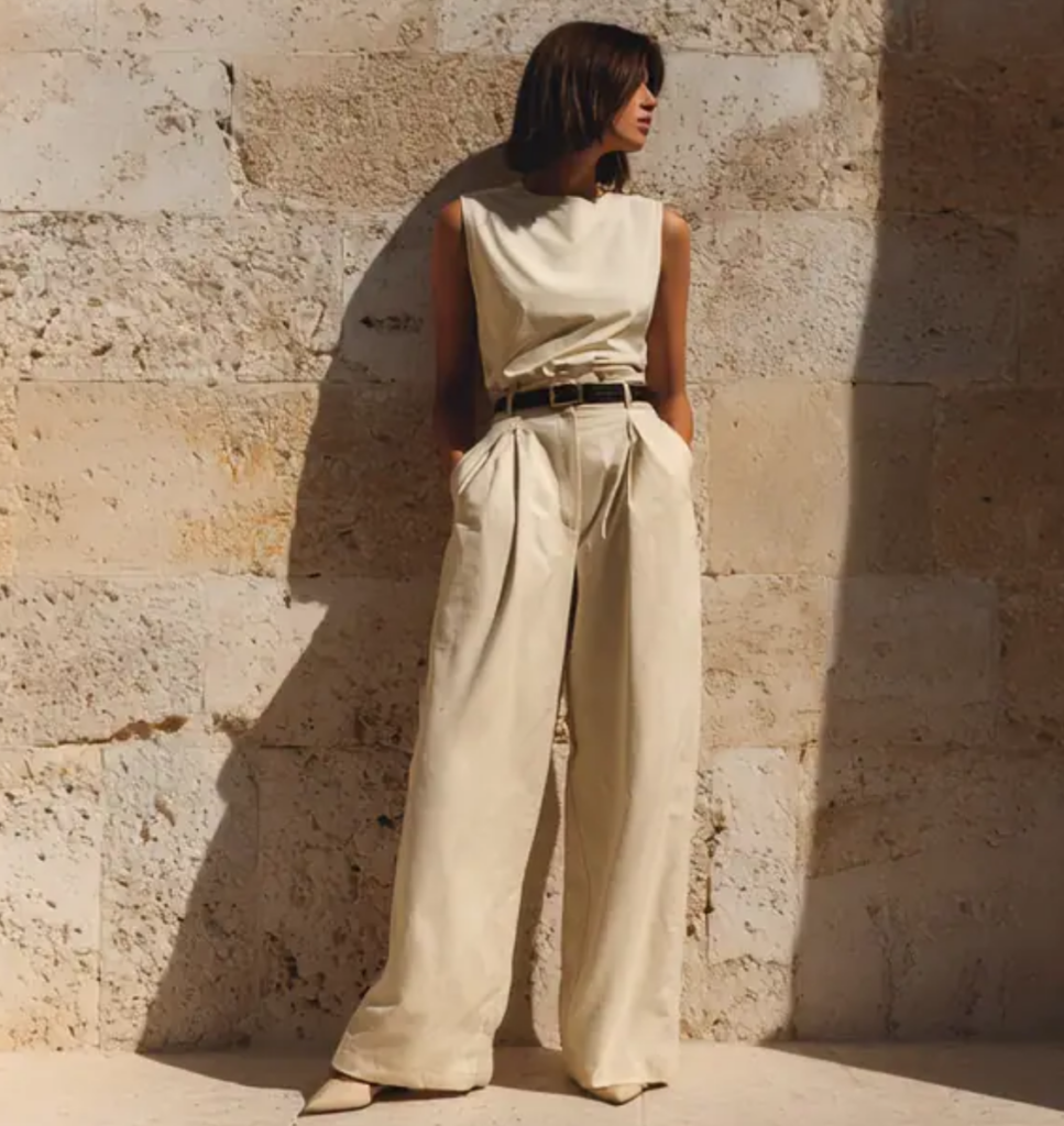 A cinematic Midjourney v7 generation of a woman in beige high-waisted trousers leaning against a sunlit stone wall.
