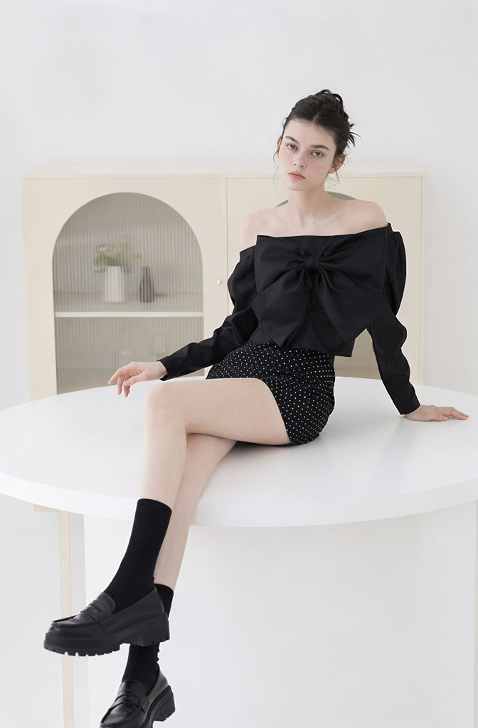 High fashion lookbook image of an AI model sitting on a white round table wearing an off the shoulder black bow top and dark patterned shorts.