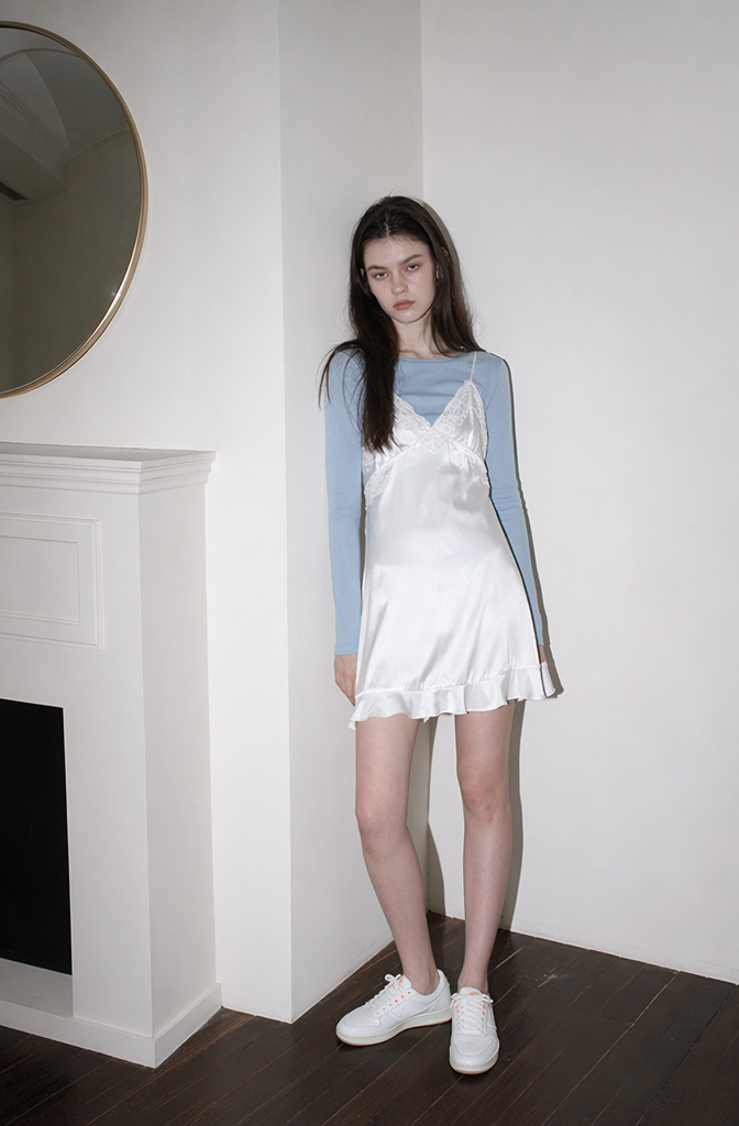 Modern fashion magazine photography style featuring an AI model in a white satin slip dress and blue shirt underlay next to a white fireplace.