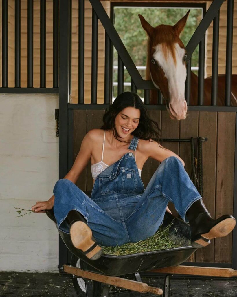 Editorial photo of celebrity Kendall Jenner wearing denim overalls sitting in a wheelbarrow with a horse background.