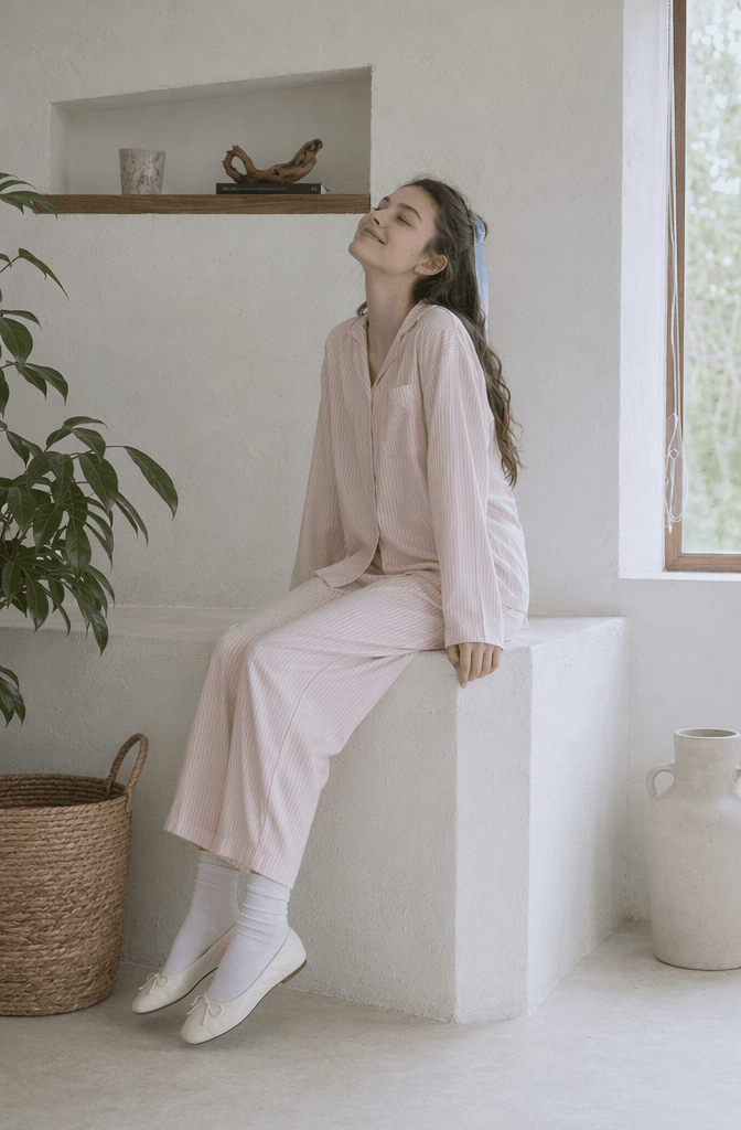 Minimalist indoor lookbook generation showing an AI fashion model in a pastel pink pajama lounge set sitting by a bright window.