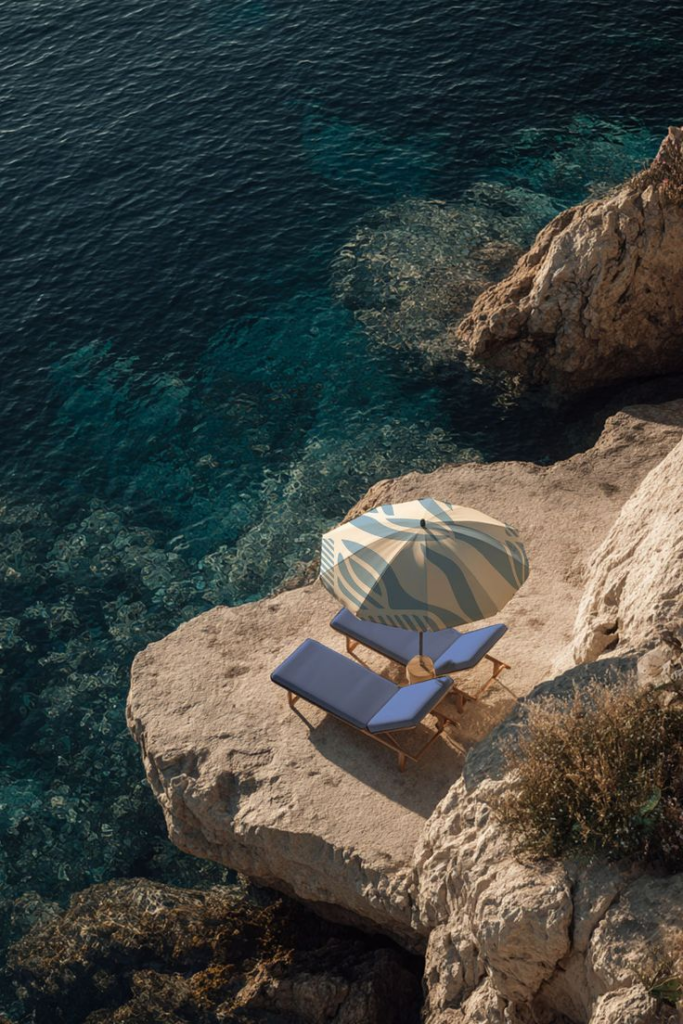 qqq – WeShop AI Blog An overhead shot of blue lounge chairs and a patterned sun umbrella perched on a rocky cliff overlooking clear turquoise Mediterranean waters.