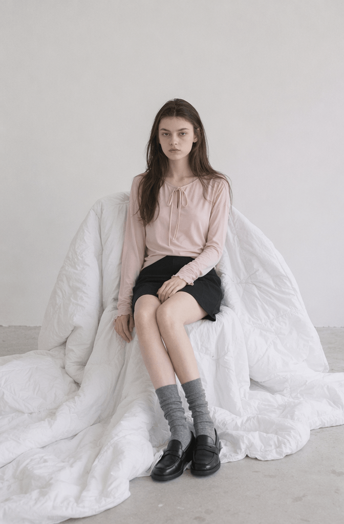 Cinematic lighting fashion lookbook photo of an AI model wearing a soft pink long-sleeve top and black skirt sitting on a white quilt blanket chair.