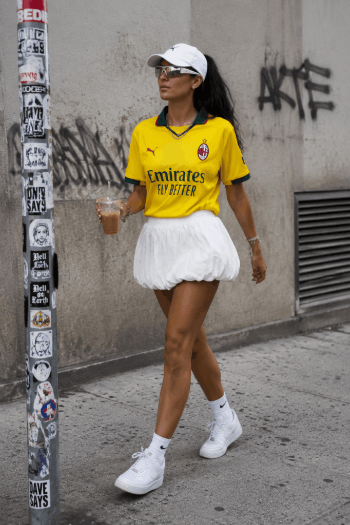 cup3 – WeShop AI Blog Street style football outfit with a yellow jersey, white skirt, sneakers, sunglasses, and an iced drink, showing a wearable World Cup fashion idea.