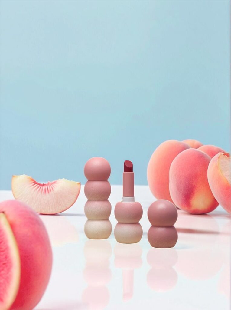 67e68c87ec14fa0372d155de_final – WeShop AI Blog AI Product Photography showing the same pink cosmetic products in a colorful peach-themed lifestyle campaign.