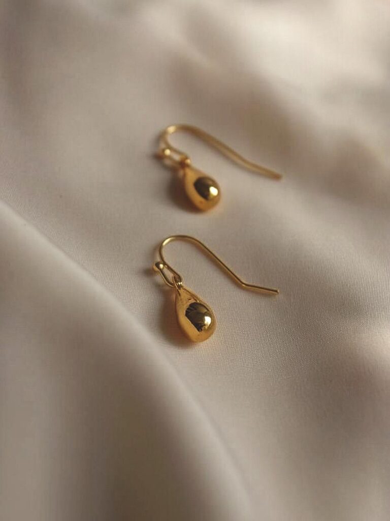 67f4a6c1394c3689928c900c_final – WeShop AI Blog Gold drop earrings on beige satin fabric after AI Relight with brighter highlights and clearer shadows.