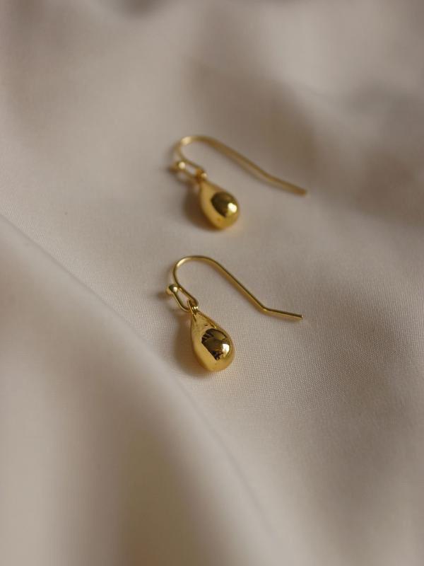 67f4a6c1394c3689928c900c_original 1 – WeShop AI Blog Gold drop earrings on beige satin fabric before AI Relight with softer, dimmer illumination.