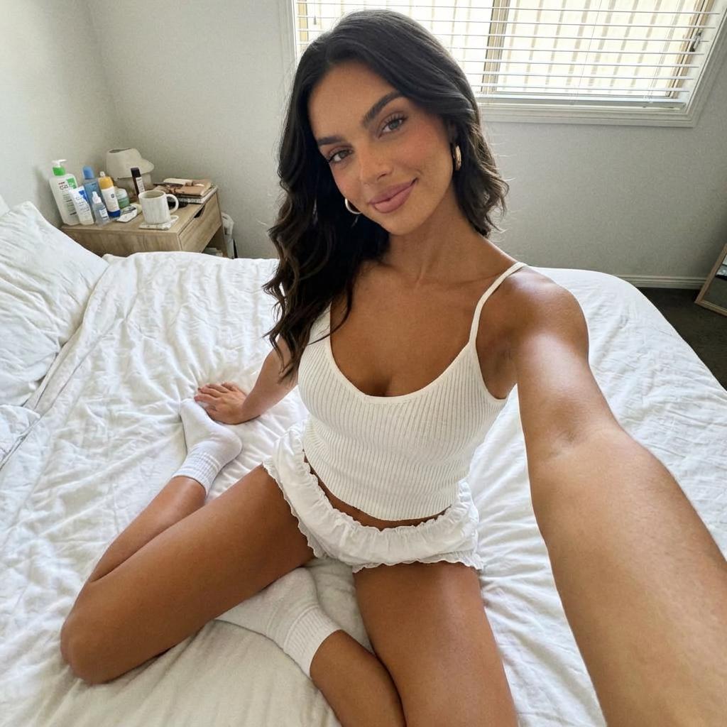 Adult woman wearing a white outfit taking a casual photo while sitting on a bed in a bright bedroom.