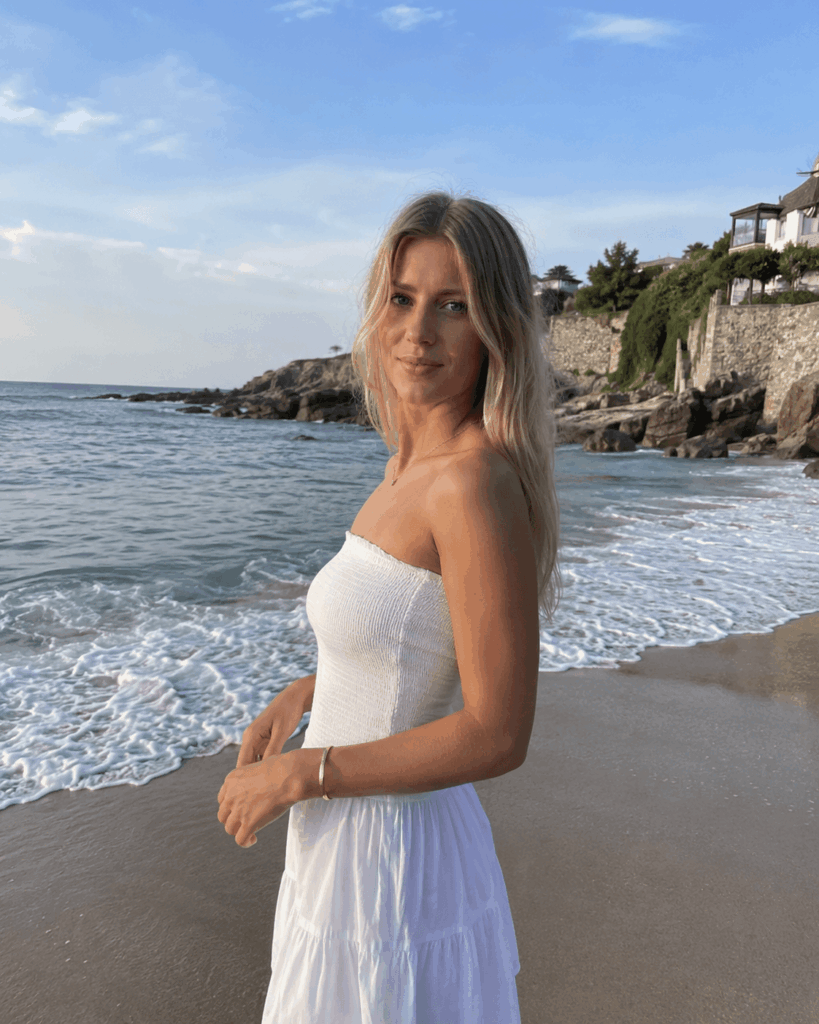 Blonde woman in a white dress standing beside the beach in natural daylight.