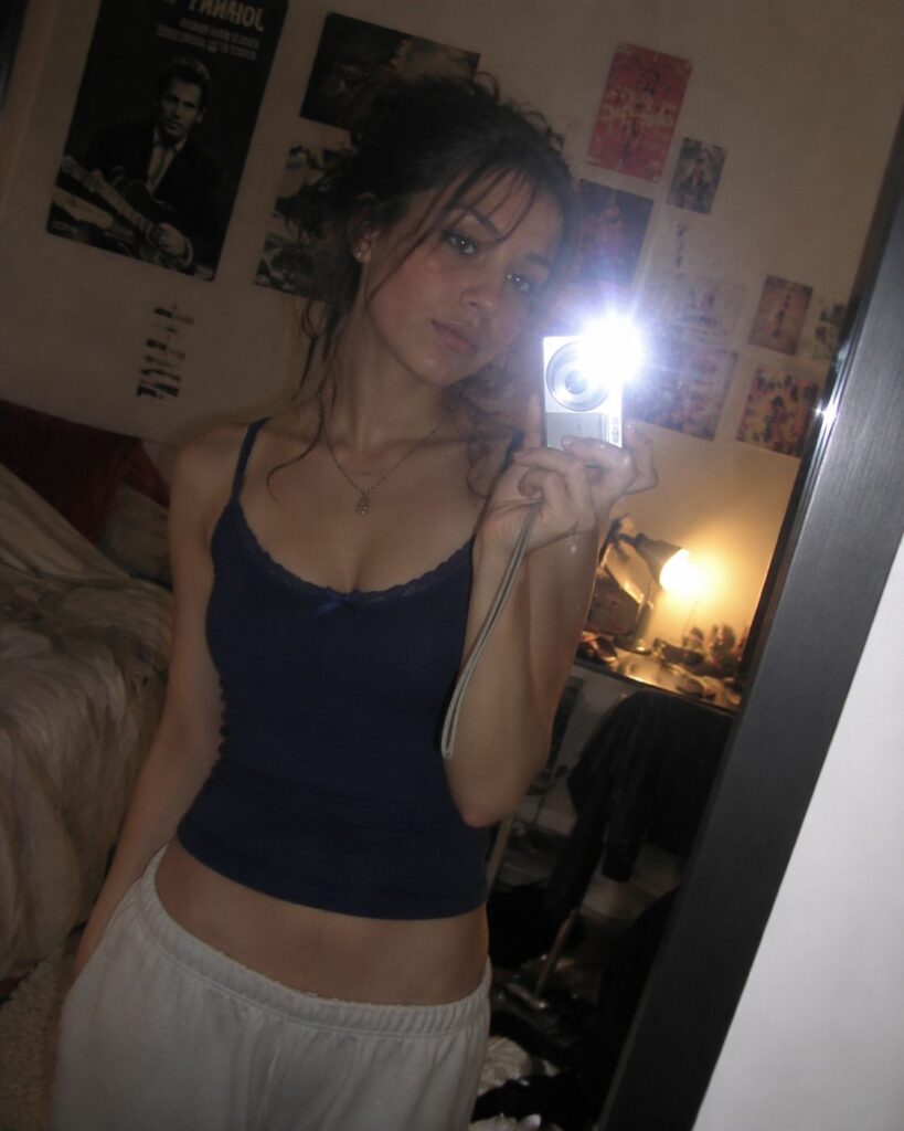 Task-126945702-1-1 – WeShop AI Blog Bedroom mirror selfie with a woman holding a compact digital camera, bright flash covering part of her face, casual clothing, wall posters, warm bedside lighting, and an authentic early-2000s personal photo mood.