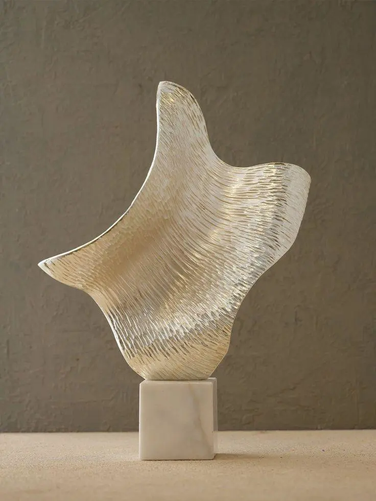Gold textured abstract sculpture displayed on a small marble base against a warm neutral wall, showing clear shape and reflective material detail.