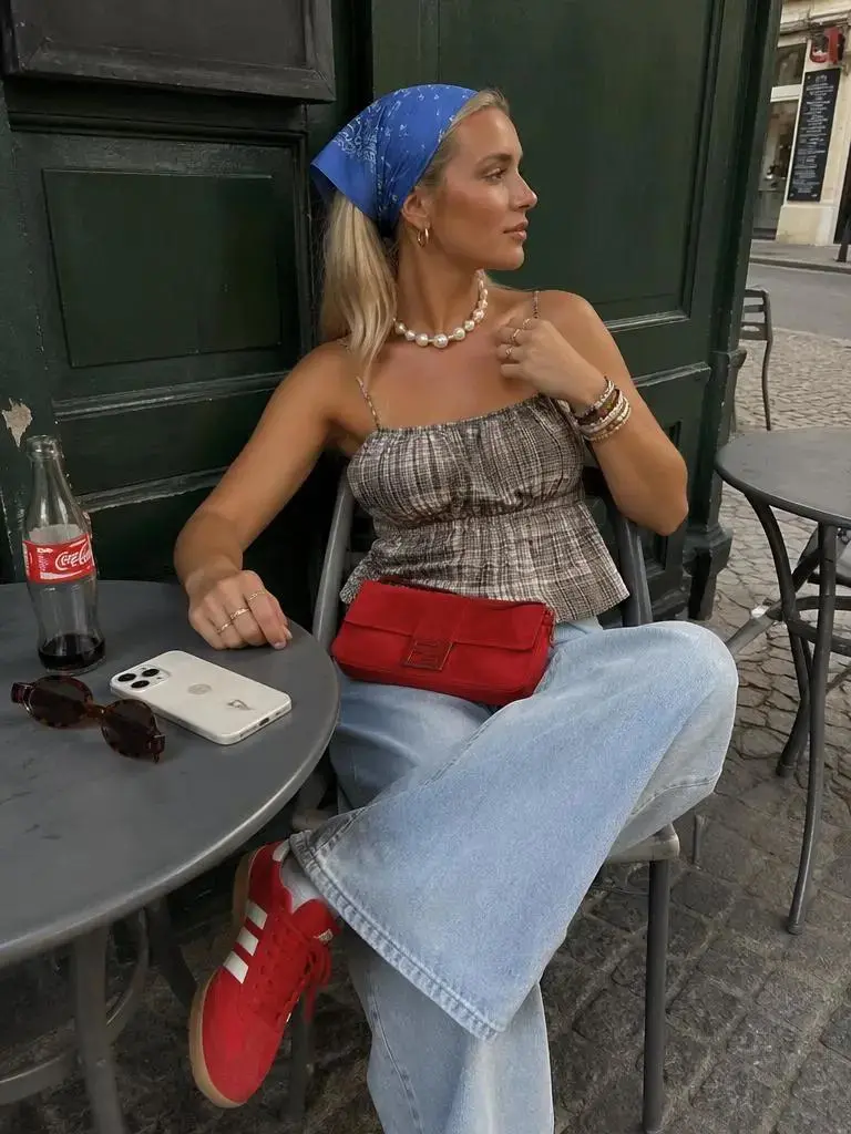 A woman wearing a blue headscarf, patterned top, jeans, and red bag sitting at an outdoor cafe.
