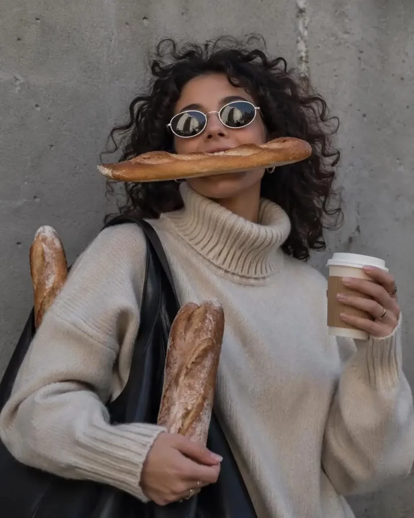 A woman with curly hair wearing sunglasses and a sweater while holding bread and coffee in a casual street photo.