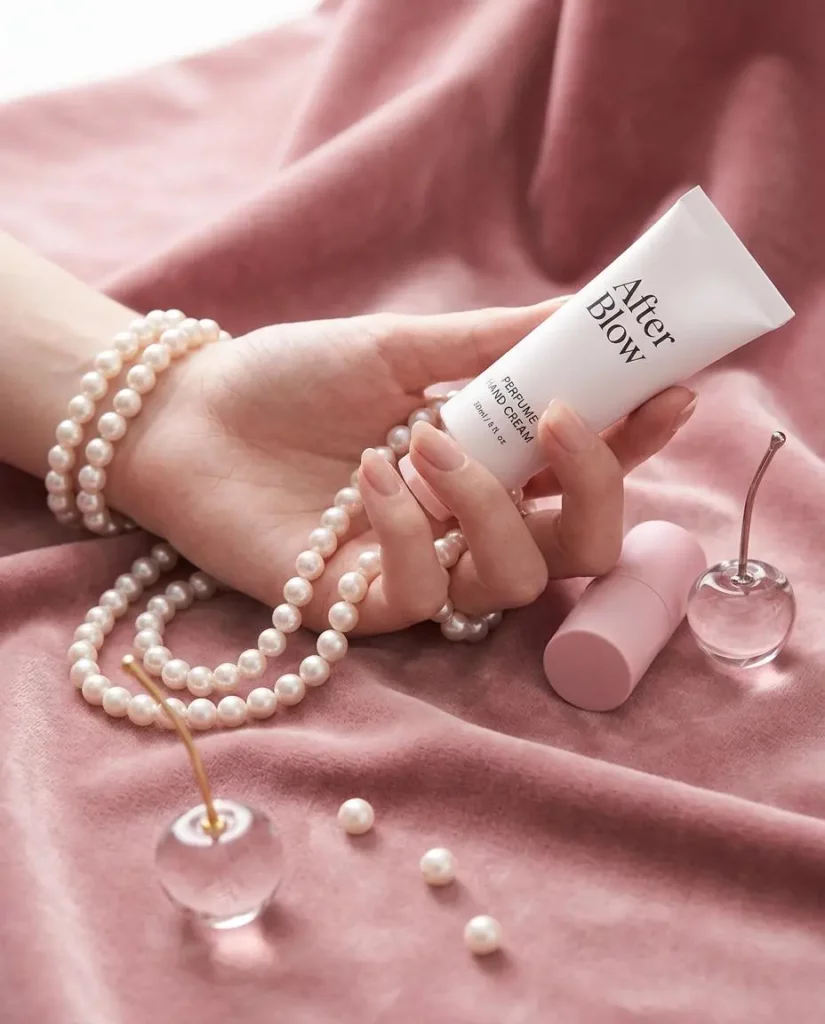 pdcd – WeShop AI Blog Hand holding a white skincare tube above pink fabric, pearls, and glass accessories in a soft beauty campaign.
