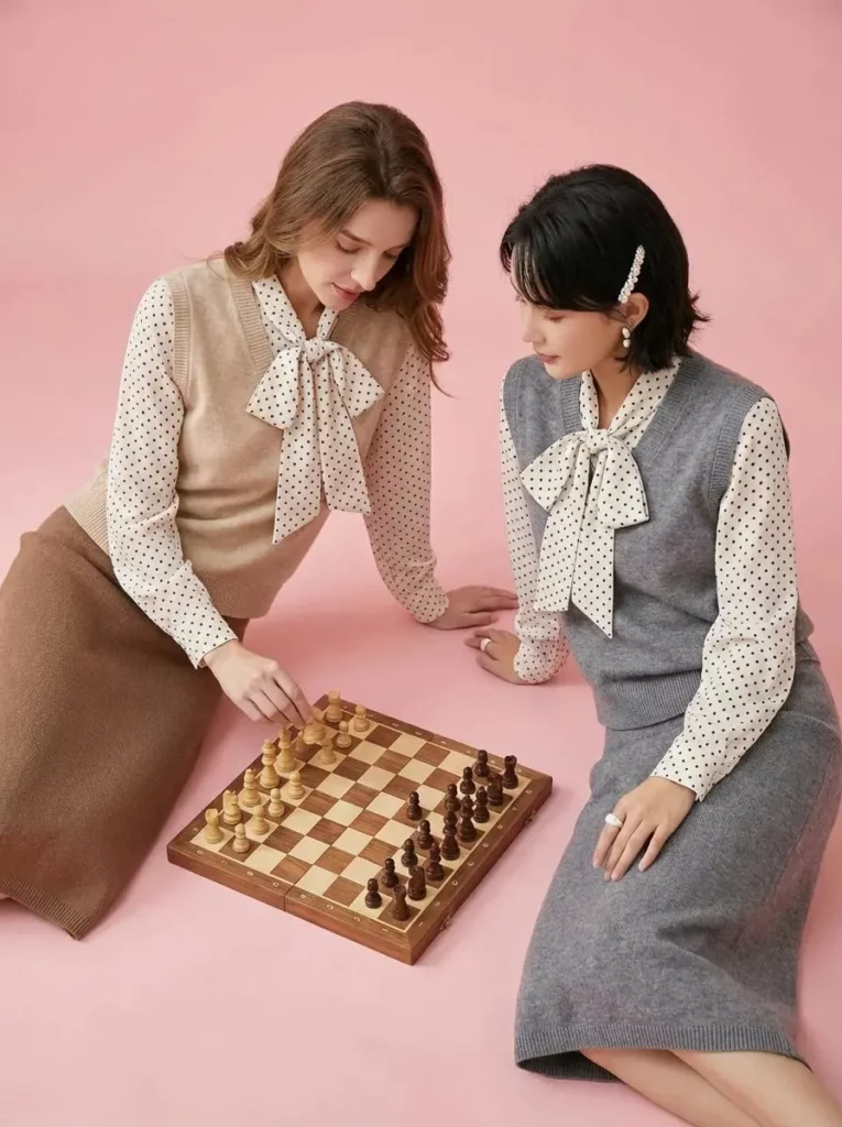poo – WeShop AI Blog Two women in beige and gray outfits seated beside a chessboard against a pink background