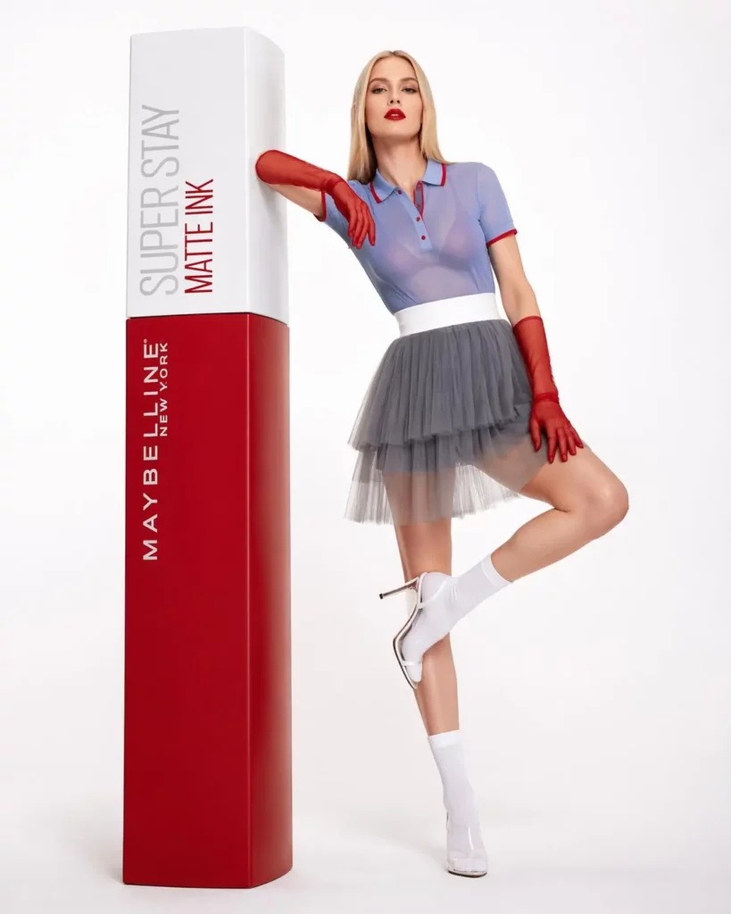 ugd – WeShop AI Blog Fashion model posing beside an oversized red lipstick in a clean commercial studio setting.